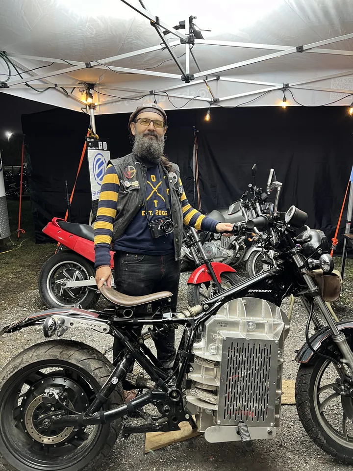 Un técnico de pie junto a una moto convertida bajo una carpa de trabajo, con el controlador y el paquete a la vista.