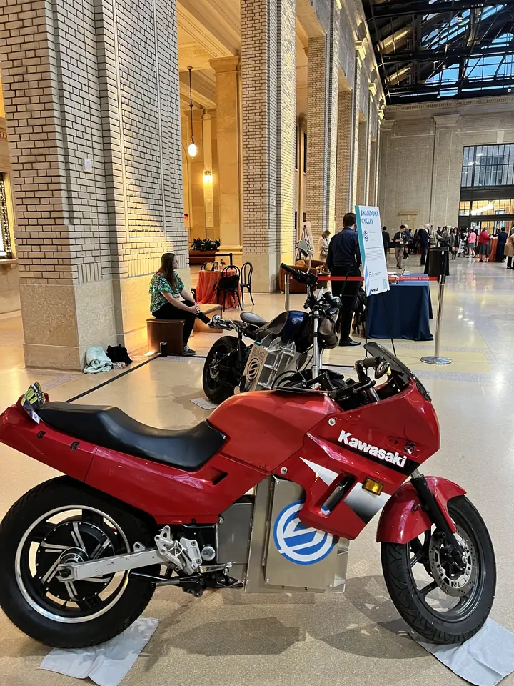La Ninja 250 eléctrica bajo techo junto a MidKnight, con el emblema Shandoka en sus baterías y el motor de buje atrás.